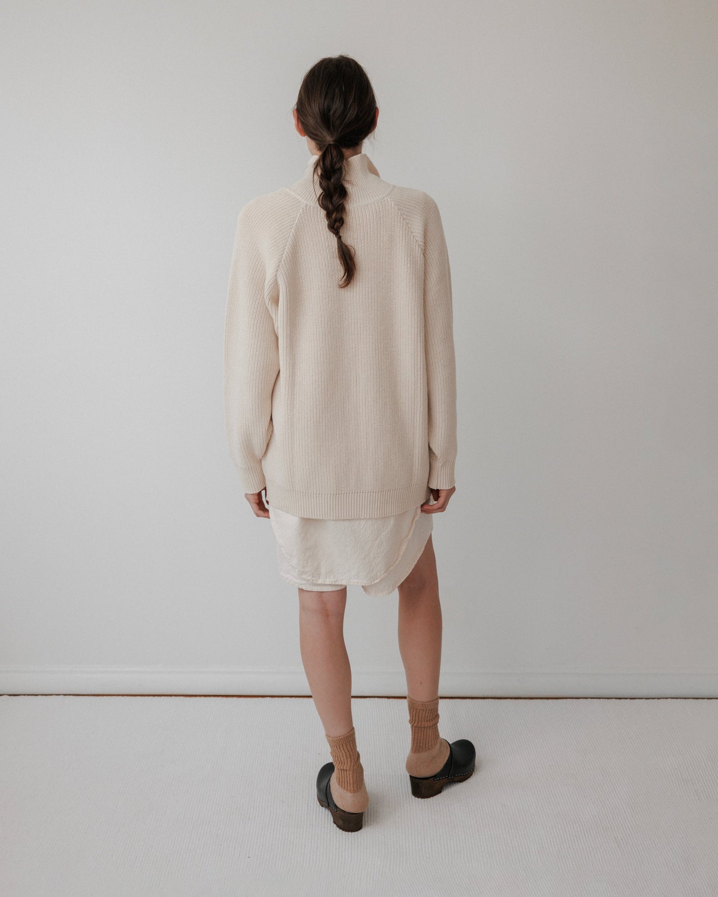A person with long, braided hair faces a white wall, wearing Wol Hide's "Collar Cardigan : Natural" over a white dress, brown socks, and dark shoes. The outfit's soft tones enhance the minimalistic setting with its plain light-colored floor.