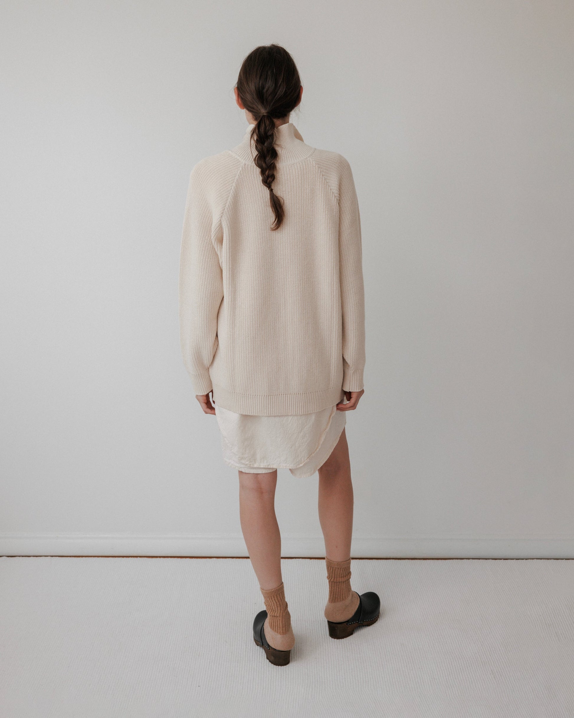 A person with long, braided hair faces a white wall, wearing Wol Hide's "Collar Cardigan : Natural" over a white dress, brown socks, and dark shoes. The outfit's soft tones enhance the minimalistic setting with its plain light-colored floor.