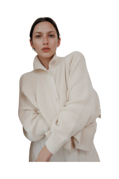 The woman wears the Wol Hide Collar Cardigan in Natural, featuring a creamy white, textured design with raglan sleeves, made in the USA. Her short hair and natural makeup enhance her confident gaze against a black background.