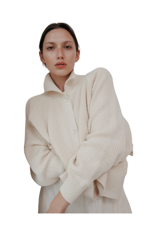 The woman wears the Wol Hide Collar Cardigan in Natural, featuring a creamy white, textured design with raglan sleeves, made in the USA. Her short hair and natural makeup enhance her confident gaze against a black background.