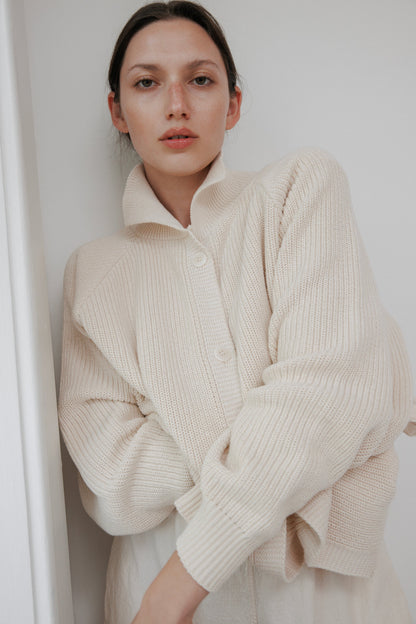 A woman stands against a white wall, arms crossed, wearing the Wol Hide Collar Cardigan: Natural. Her neutral expression and dark hair pulled back blend with the lightweight knit's minimalistic style, creating a serene look.