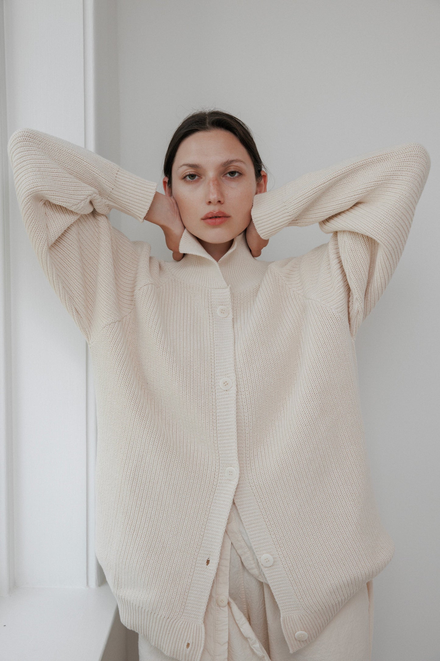 A person stands with arms raised, wearing a Wol Hide Collar Cardigan in natural. They're in a softly lit room against a pale wall, looking directly at the camera with a neutral expression.