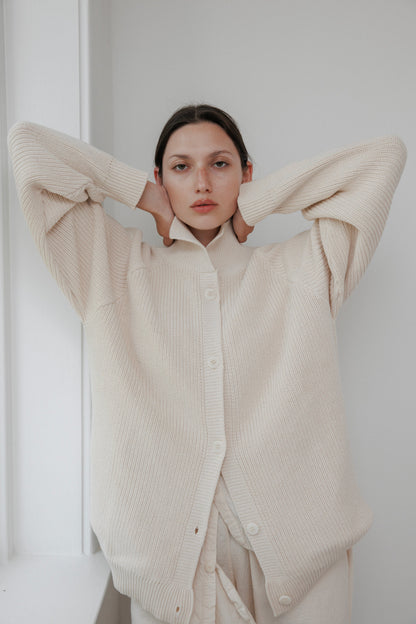 A person stands with arms raised, wearing a Wol Hide Collar Cardigan in natural. They're in a softly lit room against a pale wall, looking directly at the camera with a neutral expression.