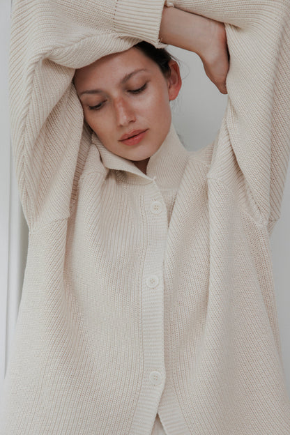 A person wearing the natural Collar Cardigan by Wol Hide, eyes closed, arms raised above their head. The neutral background creates a calm and serene mood, perfectly capturing the relaxed elegance of this USA-made garment.