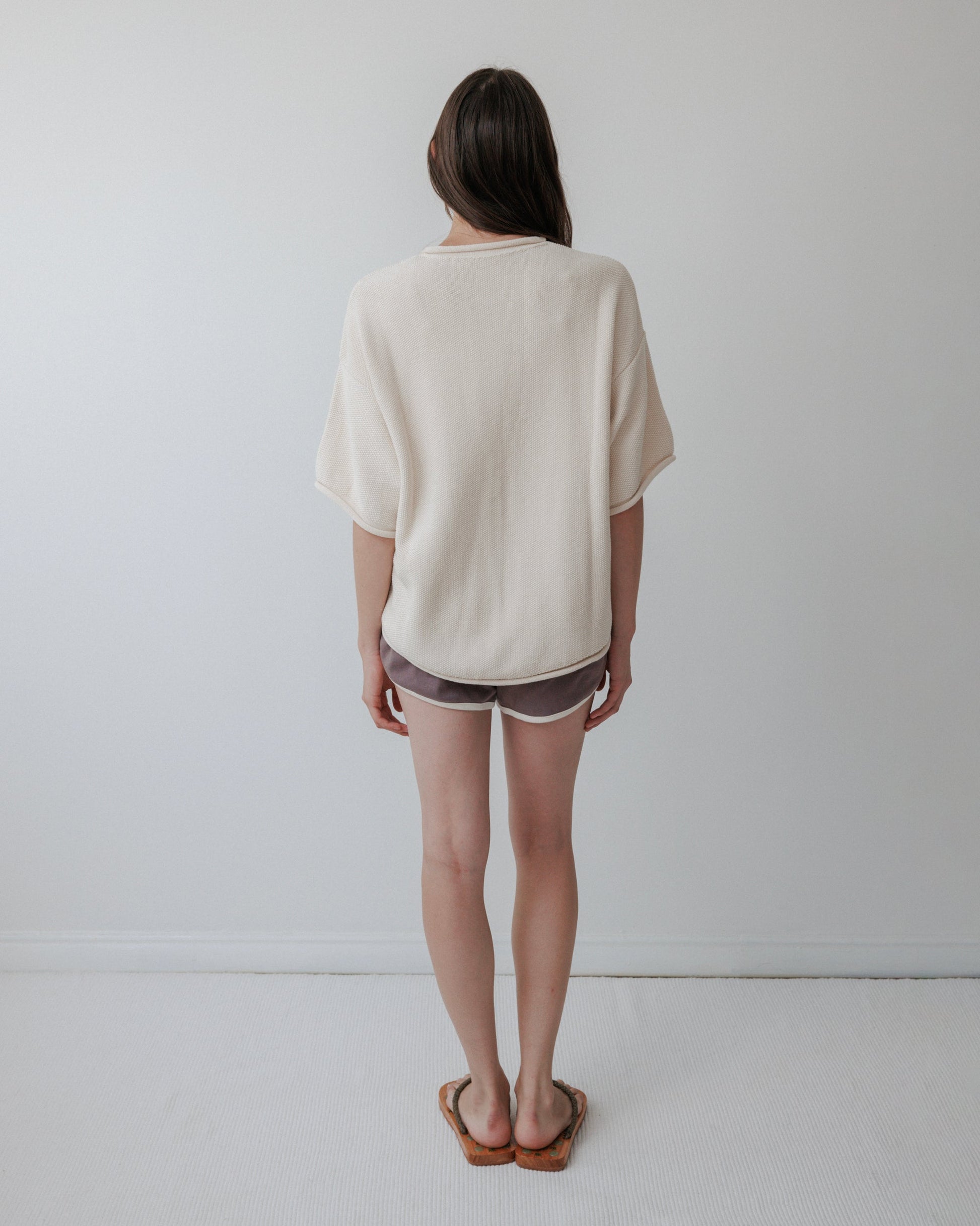 In a minimalist room, a person with long hair stands facing a white wall, wearing Wol Hide's oversized organic cotton Seed Sweater in Natural. They pair it with gray shorts featuring white trim and brown sandals, which complement the simplicity and elegance of the plain floor and walls.