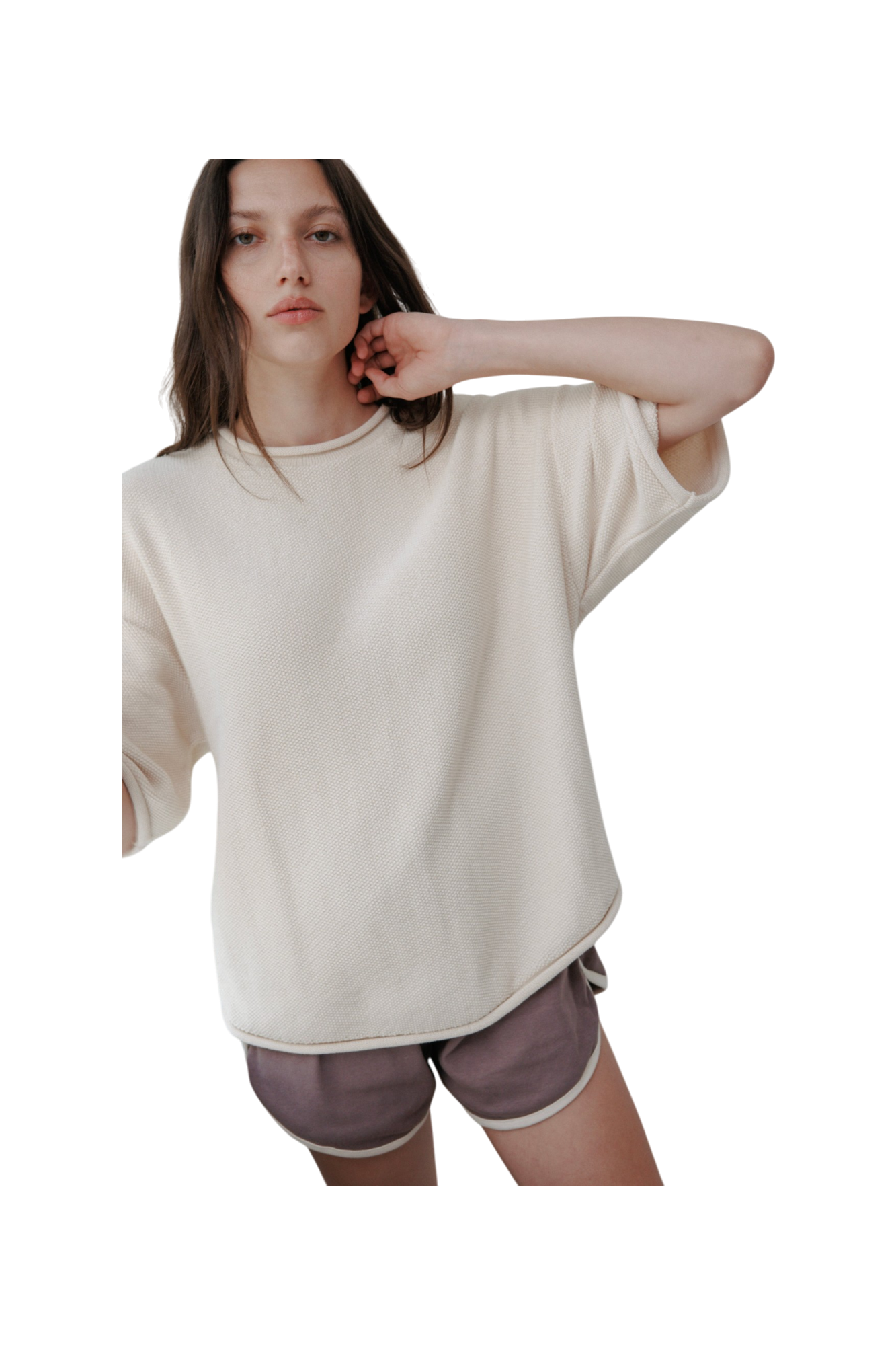 A person with long hair wears the Wol Hide Seed Sweater : Natural, an oversized organic cotton pullover, paired with purple shorts. They stand against a plain background, one arm slightly raised.