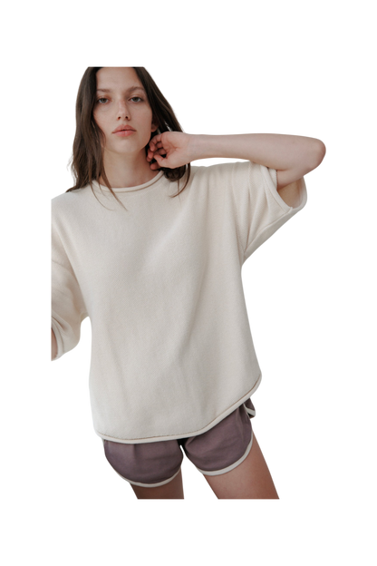 A person with long hair wears the Wol Hide Seed Sweater : Natural, an oversized organic cotton pullover, paired with purple shorts. They stand against a plain background, one arm slightly raised.