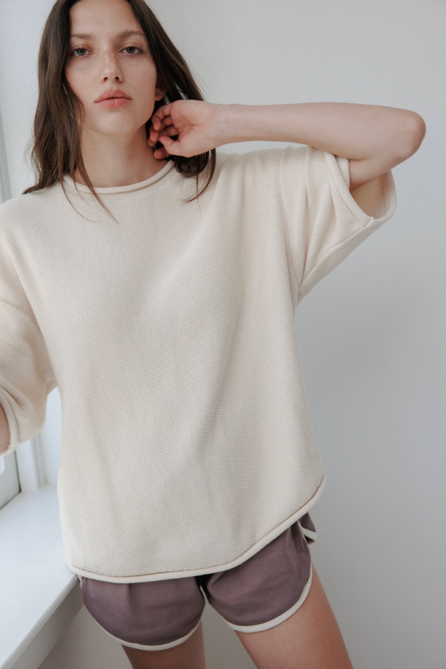 A person with long brown hair stands indoors by a window, wearing a Wol Hide Seed Sweater in natural color and mauve shorts. With their right hand touching their hair, they gaze directly at the camera.
