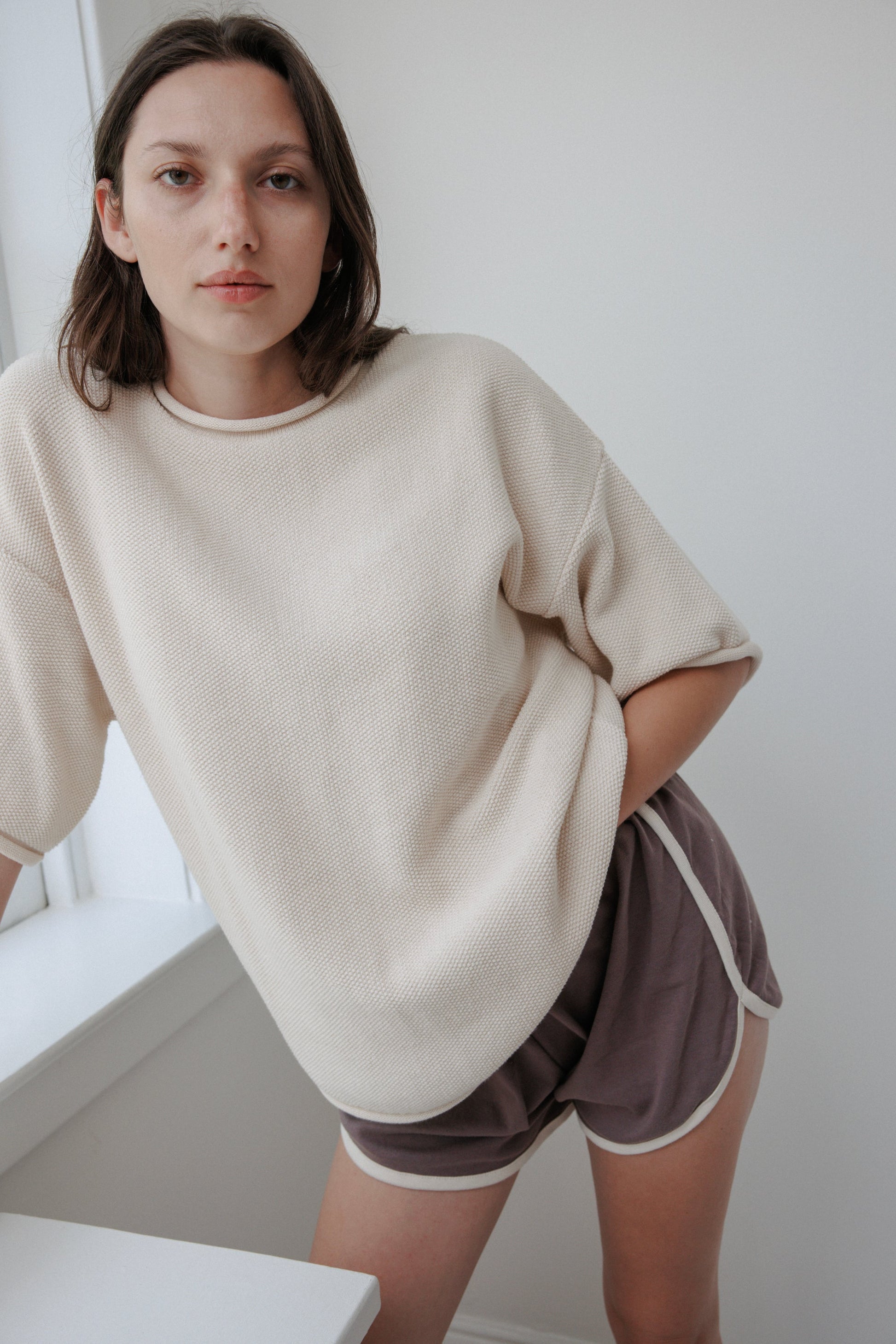 A person with shoulder-length hair leans against a softly lit windowsill, exuding effortless style and comfort in an oversized Seed Sweater : Natural by Wol Hide, made from organic cotton, paired with purple shorts featuring white trim.