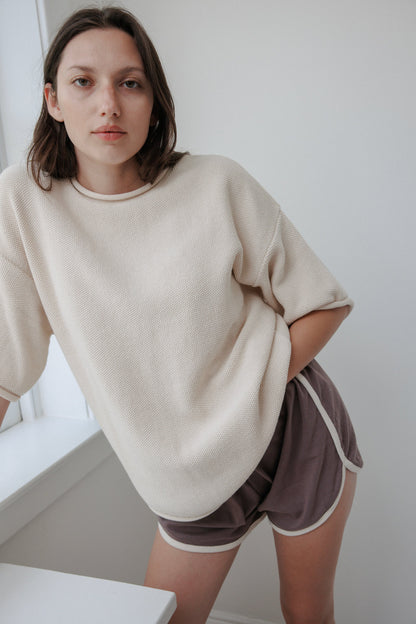 A person with shoulder-length hair leans against a softly lit windowsill, exuding effortless style and comfort in an oversized Seed Sweater : Natural by Wol Hide, made from organic cotton, paired with purple shorts featuring white trim.
