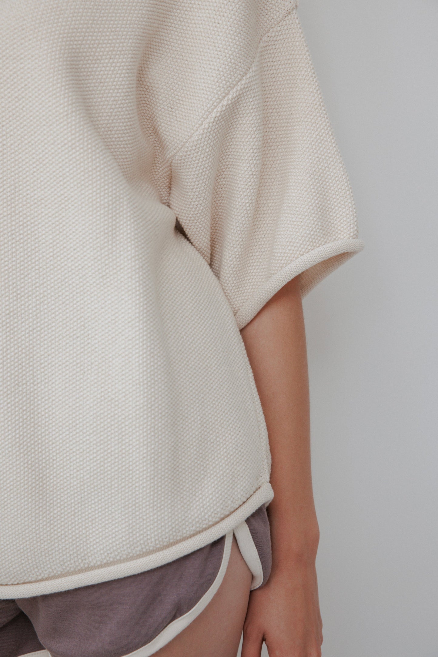 A person wearing a Seed Sweater in Natural by Wol Hide paired with mauve shorts with white trim, seen from the midsection down against a light backdrop.