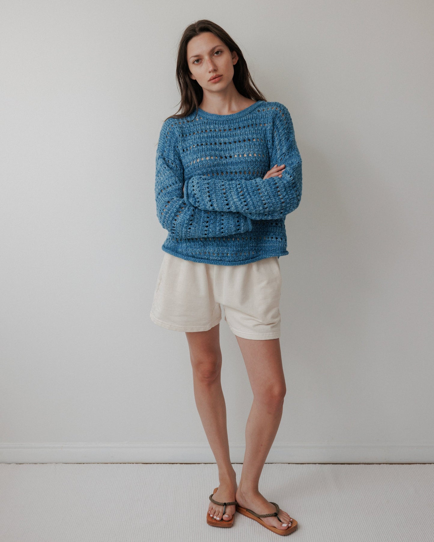 Against a plain white wall, a person wears an Open Pullover : Indigo by Wol Hide, made from hand-loomed, plant-dyed organic cotton. Their long brown hair cascades as they gaze at the camera with a neutral expression, complemented by white shorts and brown sandals.