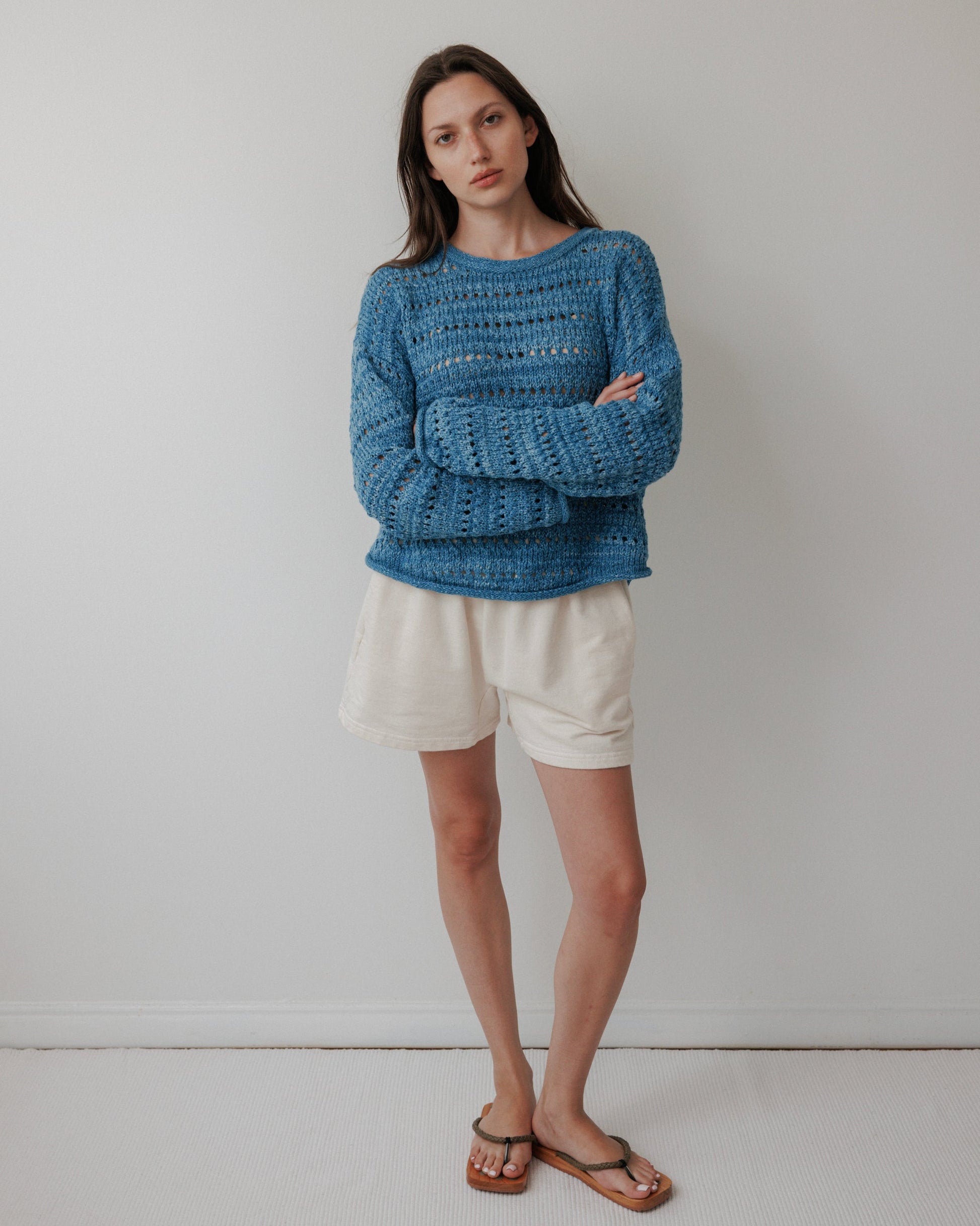 Against a plain white wall, a person wears an Open Pullover : Indigo by Wol Hide, made from hand-loomed, plant-dyed organic cotton. Their long brown hair cascades as they gaze at the camera with a neutral expression, complemented by white shorts and brown sandals.