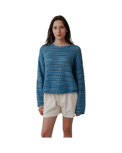 A person stands against a plain background wearing the Wol Hide Open Pullover in Indigo paired with beige shorts, looking directly at the camera.