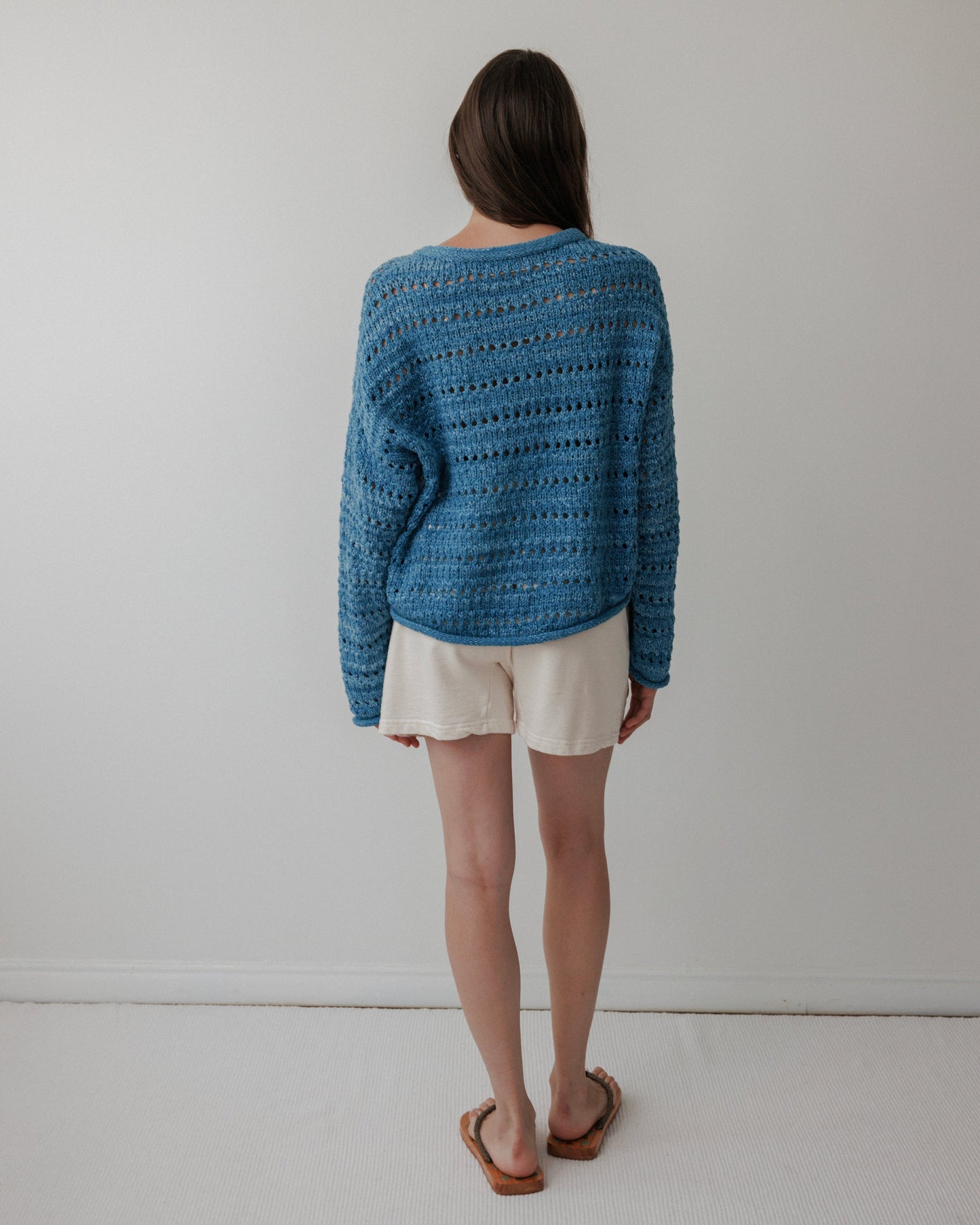 A person with long brown hair stands facing away, wearing a Wol Hide Open Pullover in Indigo, white shorts, and sandals. The background is a plain light-colored wall with a white floor.