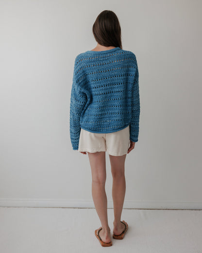 A person with long brown hair stands facing away, wearing a Wol Hide Open Pullover in Indigo, white shorts, and sandals. The background is a plain light-colored wall with a white floor.