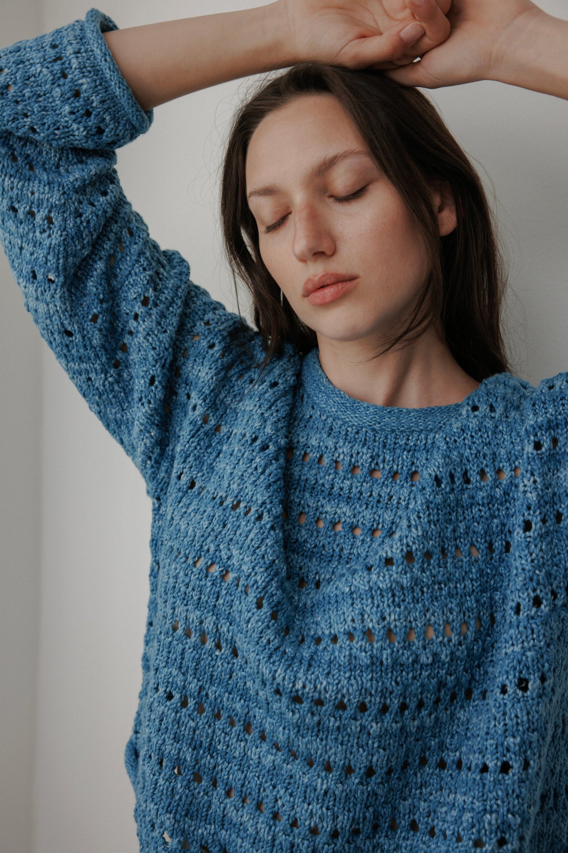 A person stands with closed eyes, arms raised, in the Wol Hide Open Pullover: Indigo. Made from plant-dyed organic cotton, the textured knit sweater enhances their relaxed demeanor as their long brown hair cascades gracefully against the plain white background.