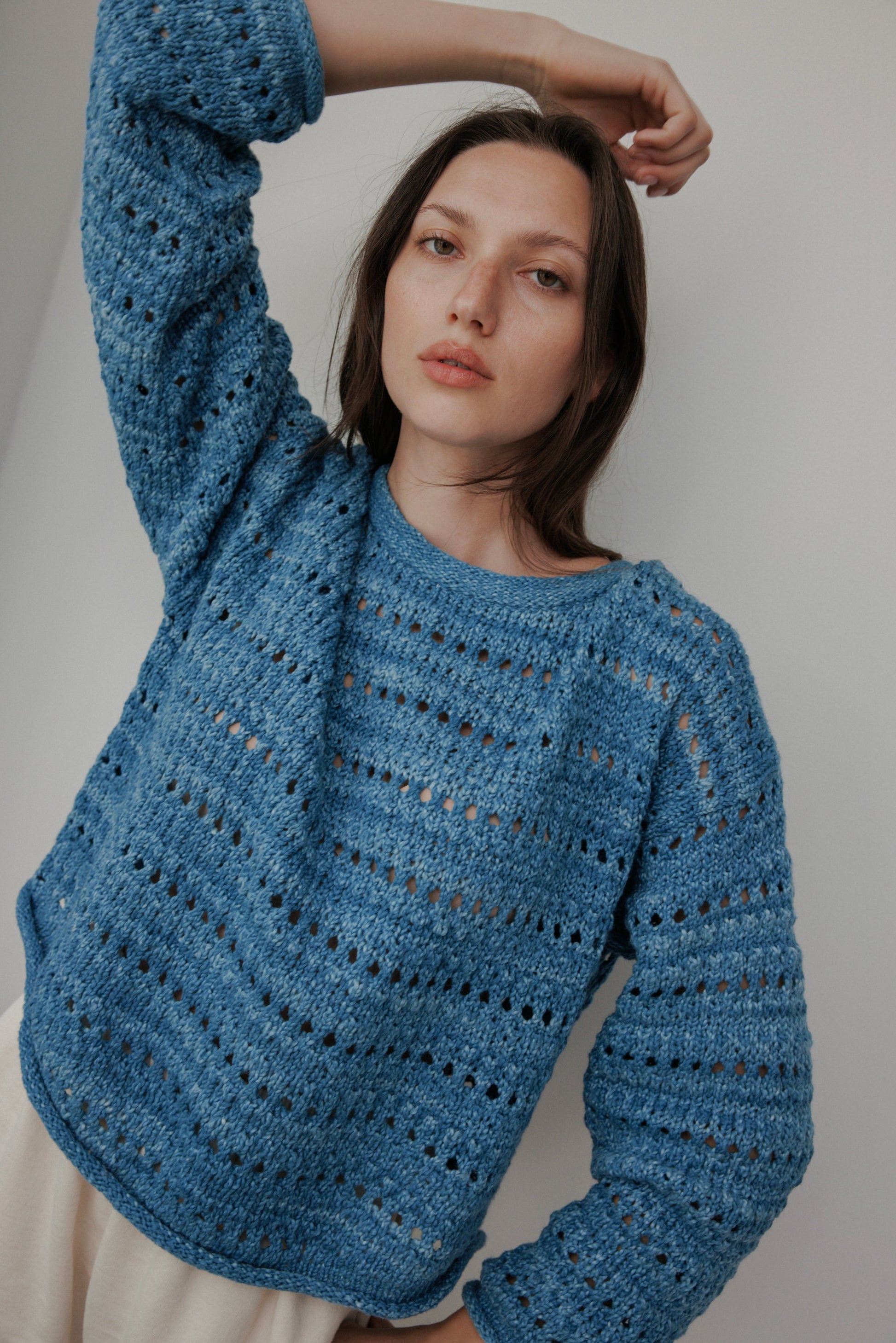 A model with long brown hair poses against a plain white background, wearing Wol Hide's Open Pullover in Indigo—a hand-loomed sweater featuring a pattern of small holes. One arm is raised behind their head as they gaze directly at the camera.