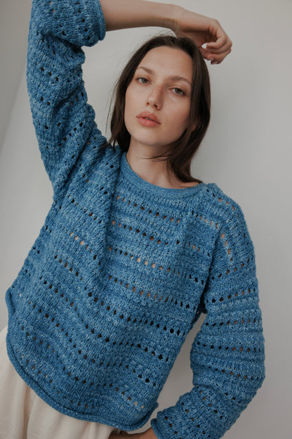 A model with long brown hair poses against a plain white background, wearing Wol Hide's Open Pullover in Indigo—a hand-loomed sweater featuring a pattern of small holes. One arm is raised behind their head as they gaze directly at the camera.