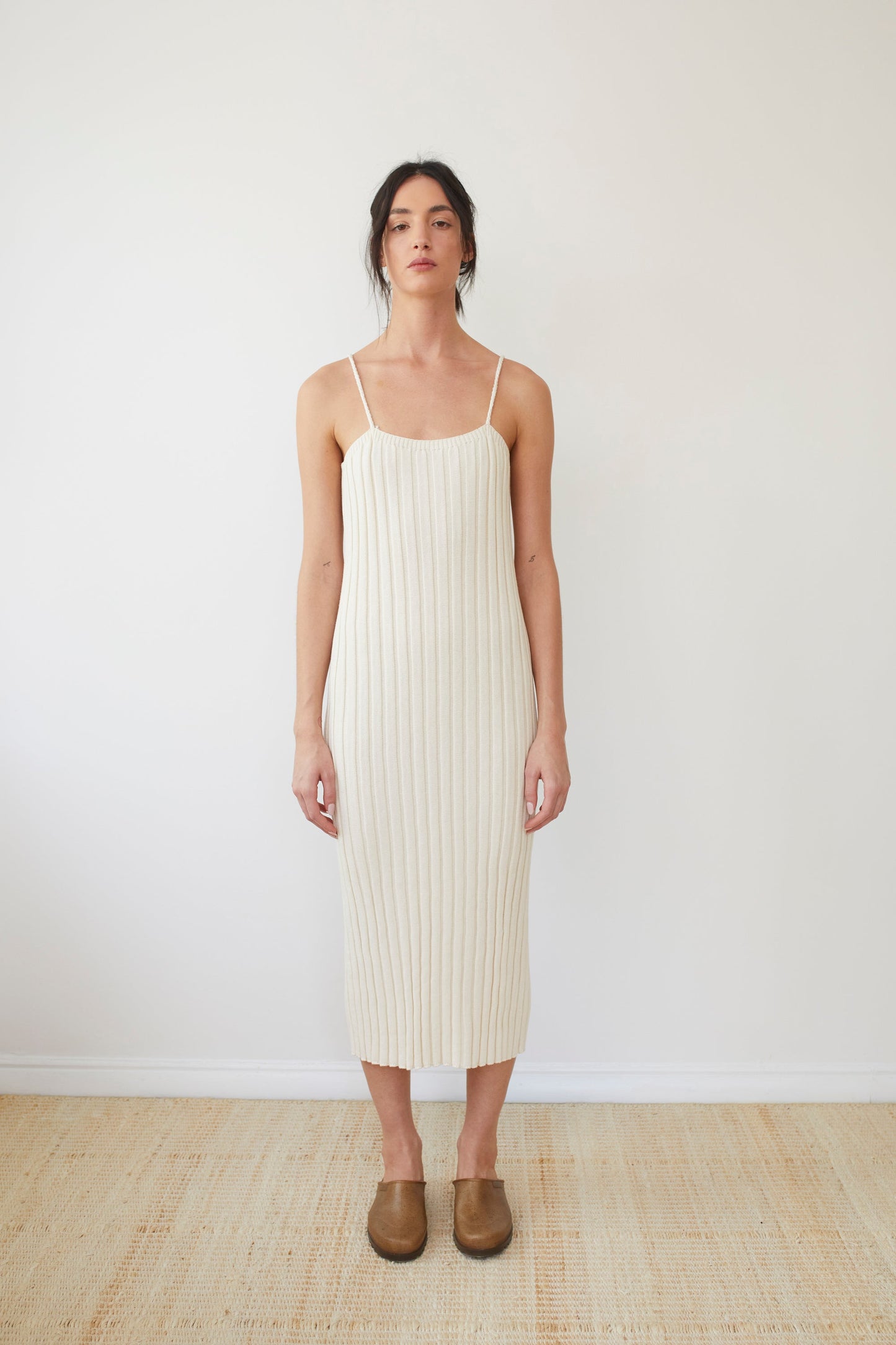 Against a plain background, a person showcases the Wol Hide Rib Dress in Ecru, an elegant sleeveless piece made from organic cotton. Their long dark hair is tied back, and they complement the look with brown slip-on shoes. The minimalistic setting is enhanced by the light-colored floor, accentuating the dress's natural beauty.