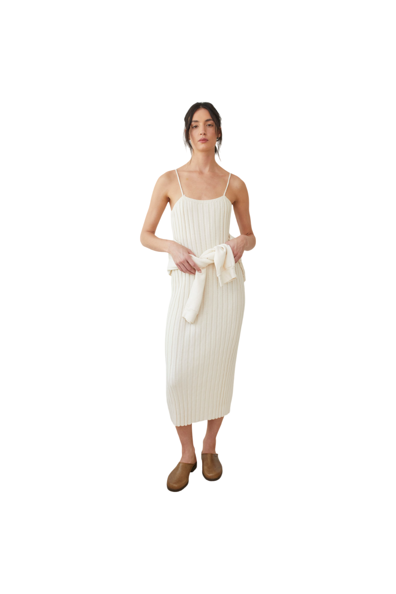 A person wearing the Rib Dress in Ecru by Wol Hide, featuring a white, sleeveless design with braided straps, is holding a cream-colored sweater tied around their waist. They have brown hair tied back and are standing against a black background, wearing brown shoes.