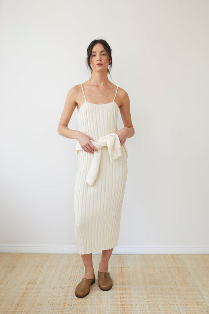 A person stands against a plain white background, wearing the Wol Hide Rib Dress in Ecru with thin straps and holding a beige sweater. They are also sporting brown slip-on shoes. The overall setting and outfit convey a minimalist style.