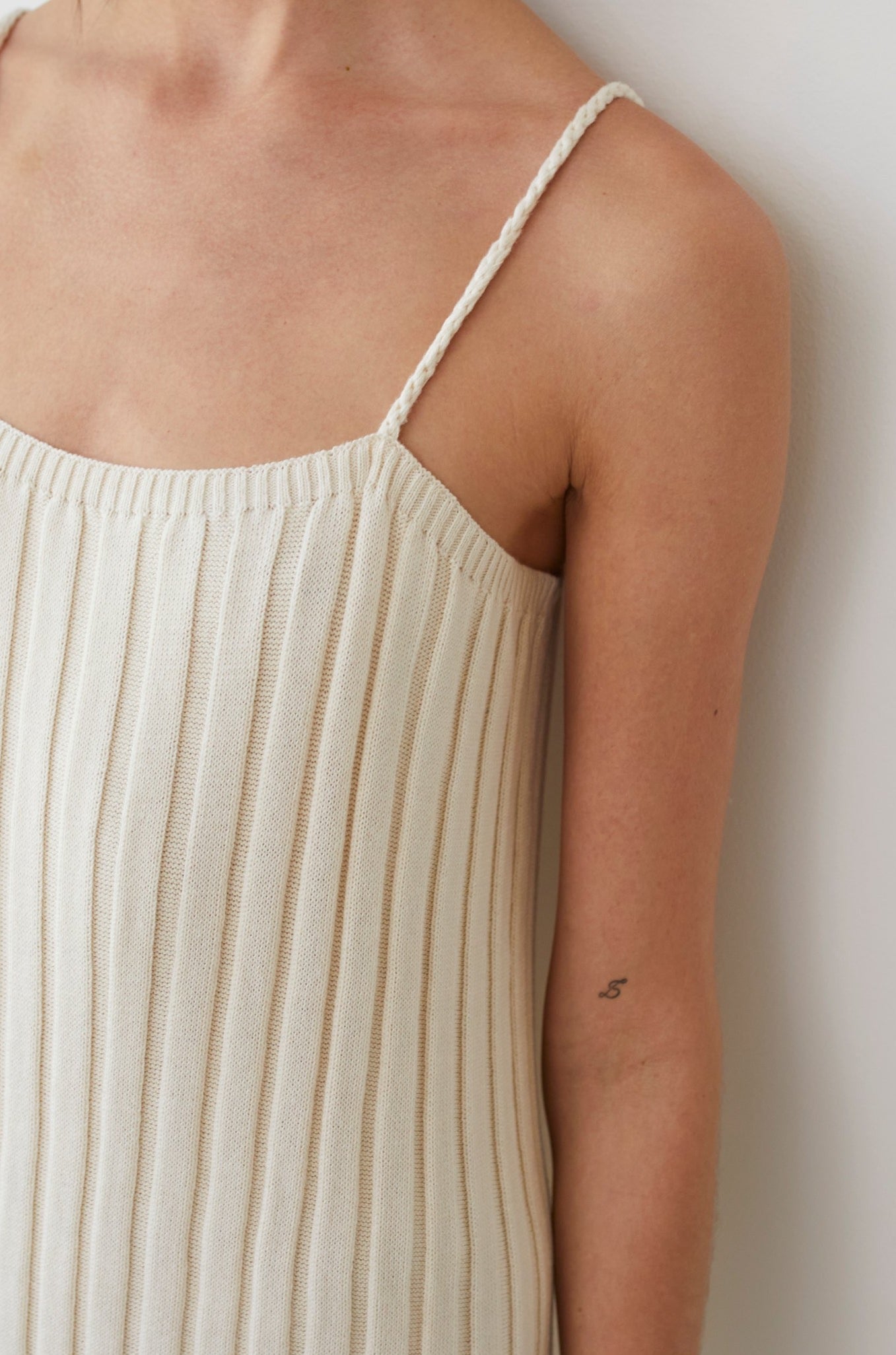 A person is wearing the Rib Dress in Ecru by Wol Hide, characterized by its sleeveless design and braided straps. A small tattoo of the number five can be seen on their arm. The background is a simple, light-colored wall.