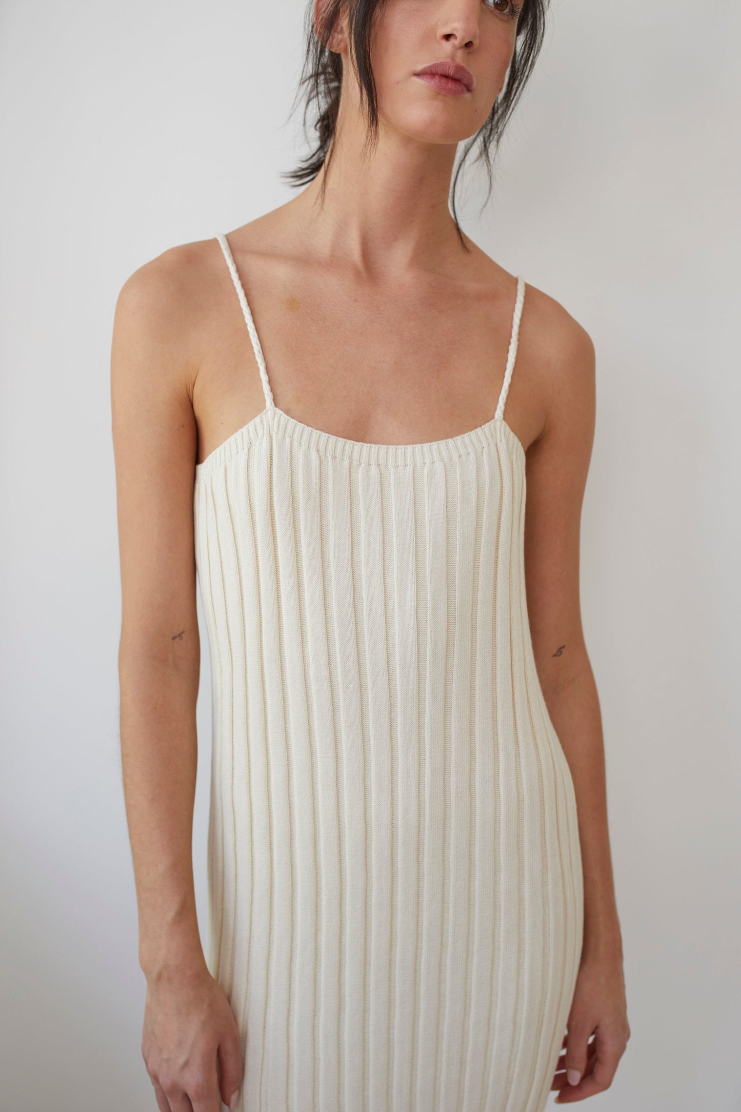 A person is standing against a plain white background, dressed in the Wol Hide Rib Dress in Ecru. The sleeveless dress is made from organic cotton, featuring thin straps, while the person's hair is tied back with a few loose strands framing their face.
