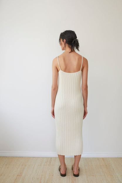 A woman stands with her back to the camera, wearing the Rib Dress in Ecru by Wol Hide. The long, sleeveless dress features braided straps and is paired with brown sandals, as she stands against a plain, light-colored wall on a beige carpeted floor.