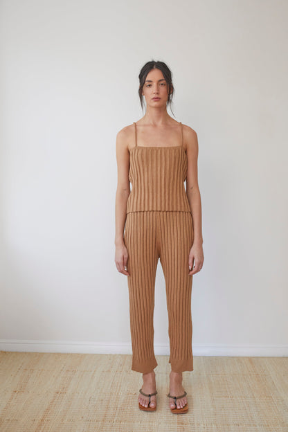 In a minimalist room, a person wears Wol Hide's Organic Cotton Rib Cami in Toffee with matching pants and strappy sandals with braided straps. Their hair is styled in a loose updo, and the floor features a woven mat.