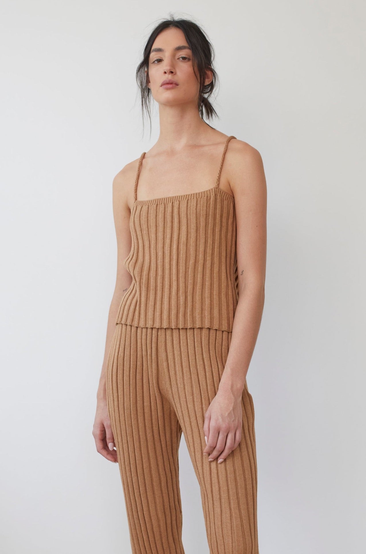 Against a white background, a person with long, dark hair in a loose updo wears Wol Hide's Organic Cotton Rib Cami in Toffee and matching pants. The outfit features thin braided shoulder straps and a textured design.