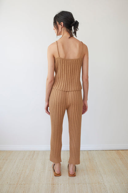 A person with dark hair tied back stands with their back to the camera, wearing Wol Hide's Organic Cotton Rib Cami in Toffee and matching pants. They're barefoot on a light-colored woven surface, showcasing an outfit made from luxurious organic cotton.