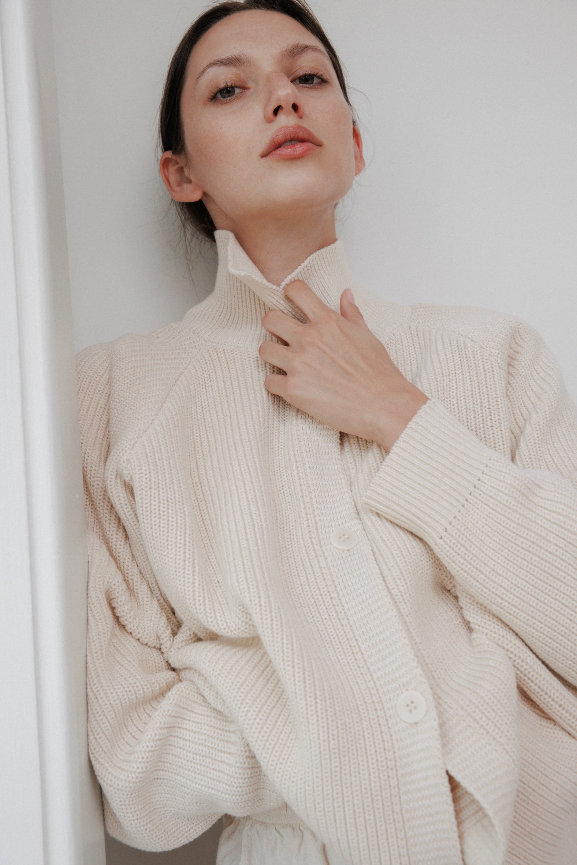 A person relaxes in a Wol Hide Collar Cardigan: Natural, gently holding the high collar with one hand. They're standing against a white background, looking upwards and exuding effortless style.