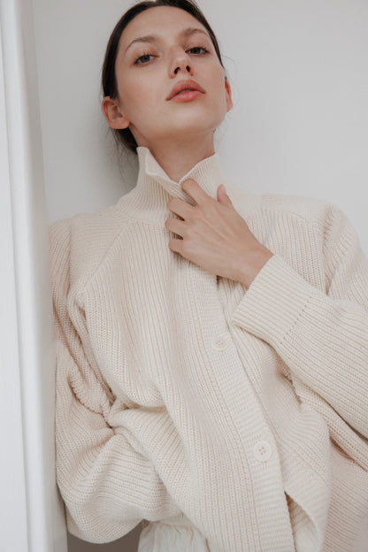 A person relaxes in a Wol Hide Collar Cardigan: Natural, gently holding the high collar with one hand. They're standing against a white background, looking upwards and exuding effortless style.