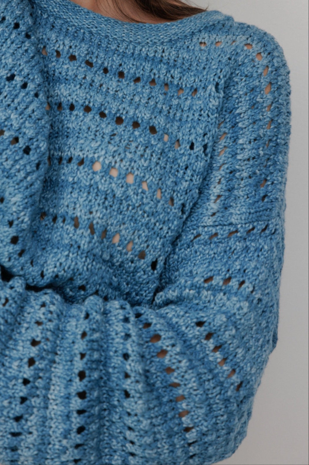 A person wears the Wol Hide Open Pullover in Indigo, featuring a textured pattern. Made from organic cotton, this hand-loomed sweater has long sleeves and offers a loose, cozy look. The background is plain light gray.