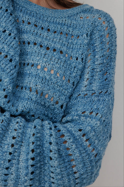 A person wears the Wol Hide Open Pullover in Indigo, featuring a textured pattern. Made from organic cotton, this hand-loomed sweater has long sleeves and offers a loose, cozy look. The background is plain light gray.