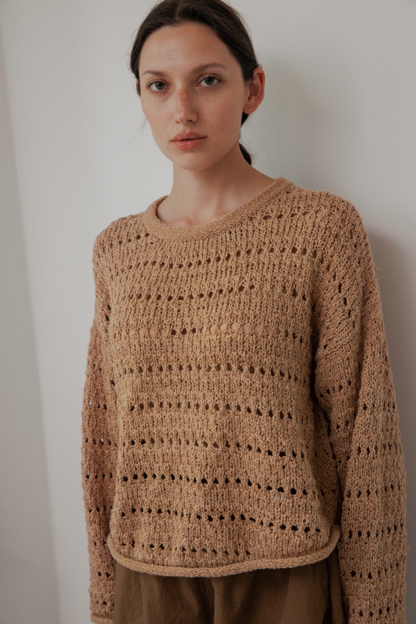 A person with long dark hair wears a Wol Hide "Open Pullover: Tannin," an artisan-crafted tan knit sweater made from organic cotton. Standing against a light wall, the hand-loomed sweater has small hole patterns that create subtle stripes. The person gazes directly at the camera with a neutral expression.