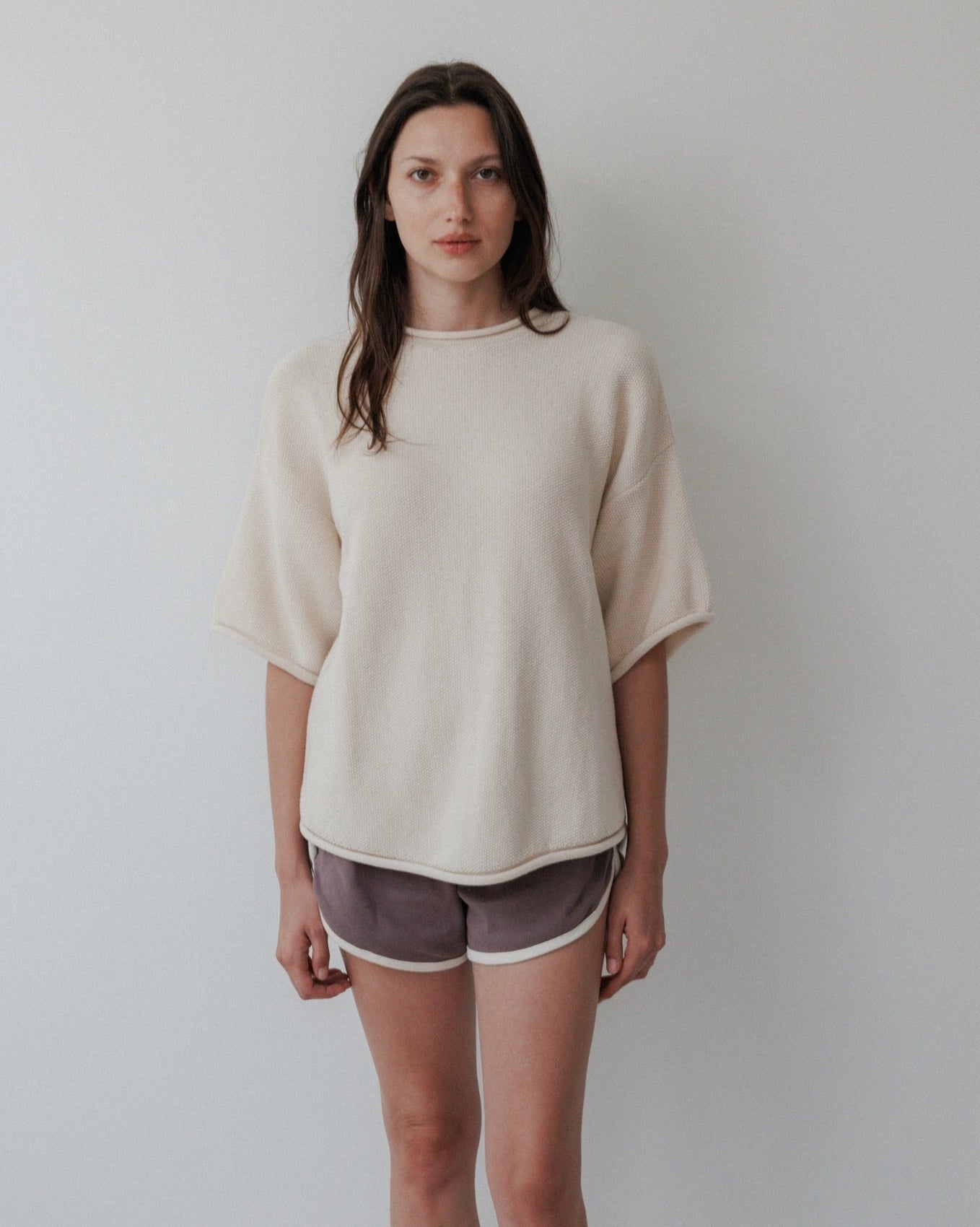 A woman with long brown hair wears the Wol Hide Seed Sweater in Natural with dark gray shorts, standing against a plain white background.