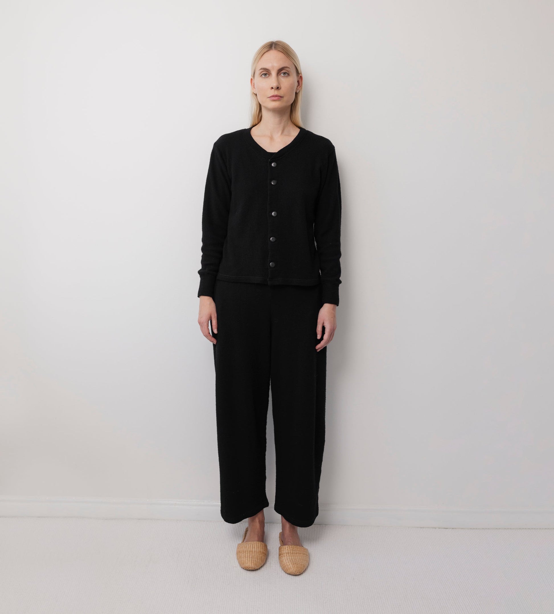 A woman with blonde hair stands against a white wall, wearing Wol Hide’s Wool Wide Leg: Onyx pants and a black button-up sweater, paired with beige woven slippers. She faces forward with a neutral expression and relaxed posture.