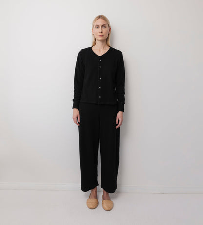 A person with light hair stands against a plain white wall, wearing the Wol Hide Wool Liner in Onyx, paired with black wide-leg pants and beige woven slippers. They face forward with a neutral expression.