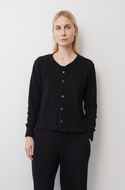 A person with long blonde hair, wearing the Wol Hide Wool Liner in Onyx and matching black pants, stands against a plain white wall and looks directly at the camera with a neutral expression.