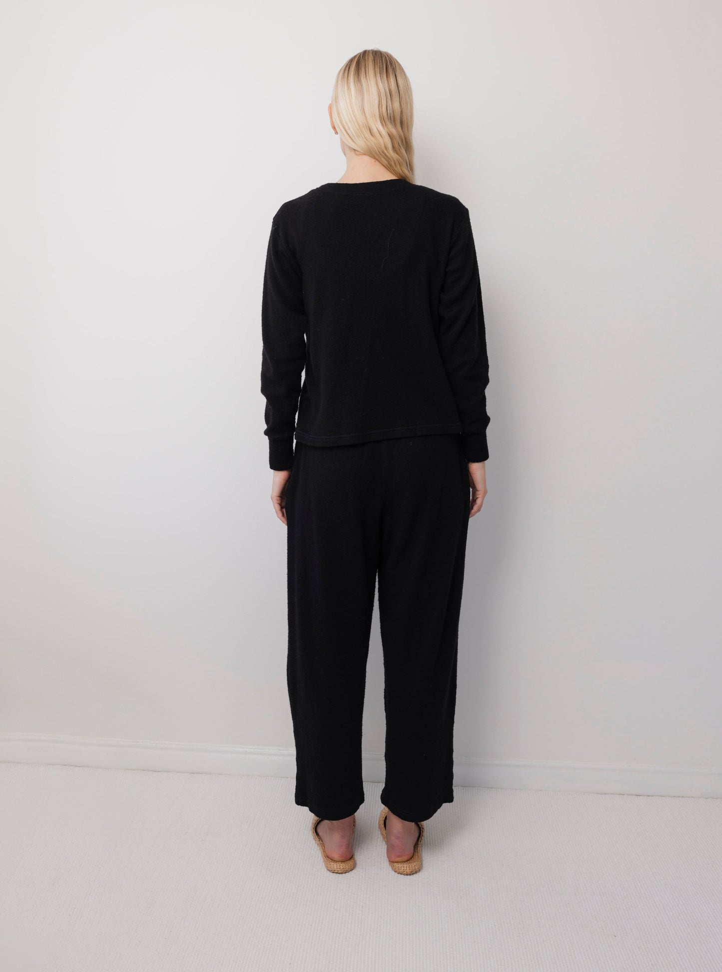 A person with long blonde hair stands facing a white wall, wearing Wol Hide’s Wool Liner in Onyx, styled with black pants and beige sandals. The outfit features corozo buttons, set against a minimal white floor and wall backdrop.