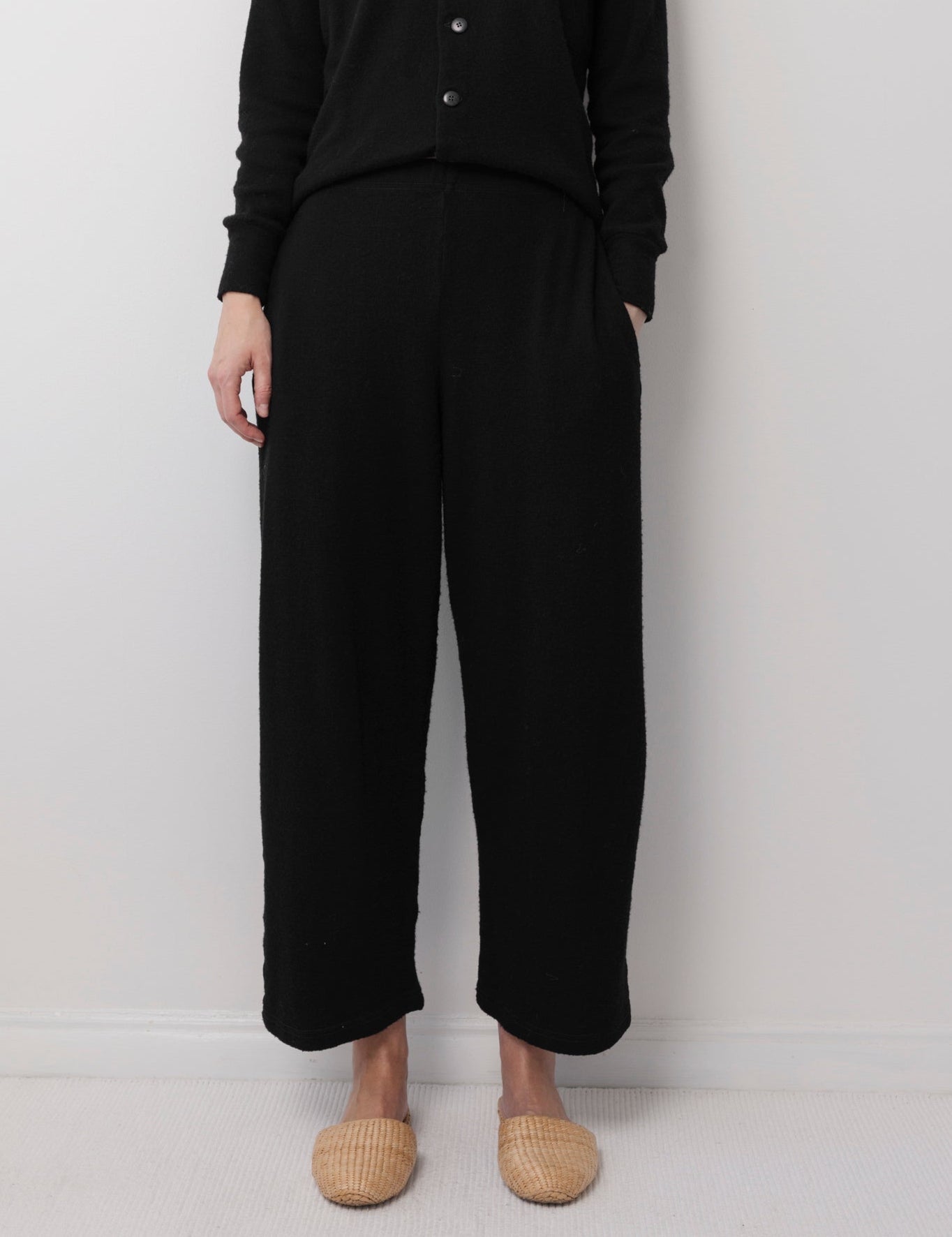A person stands against a light wall wearing Wol Hide’s Wool Wide Leg: Onyx pants with a black button-up top and beige woven slippers. Their hands are at their sides, and their head is not visible in the image.