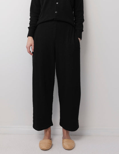 A person stands against a light wall wearing Wol Hide’s Wool Wide Leg: Onyx pants with a black button-up top and beige woven slippers. Their hands are at their sides, and their head is not visible in the image.