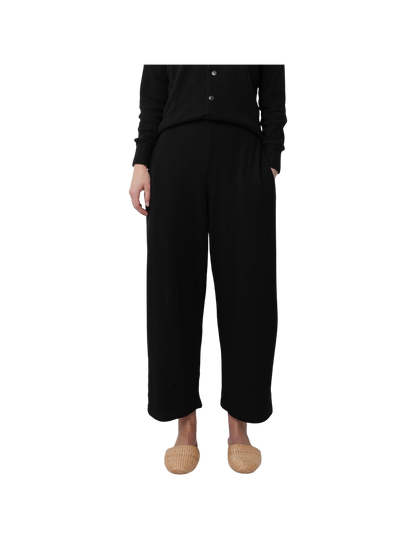 A person models the Wol Hide Wool Wide Leg pants in Onyx with a matching black buttoned top and light woven slippers, standing against a plain white background. Only their lower body is visible.