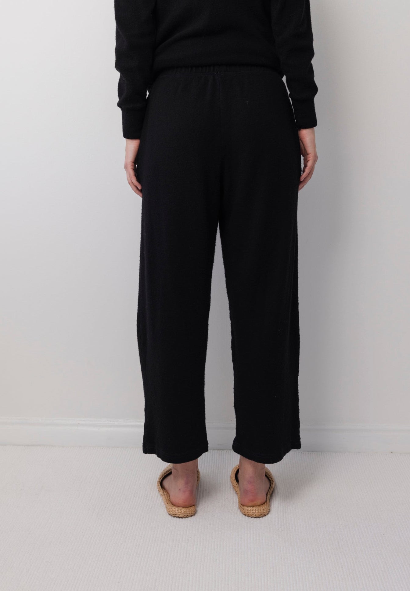 A person stands with their back to the camera, wearing the Wol Hide Wool Wide Leg: Onyx pants, a long-sleeve black top, and flat woven sandals against a plain white wall and light-colored floor.