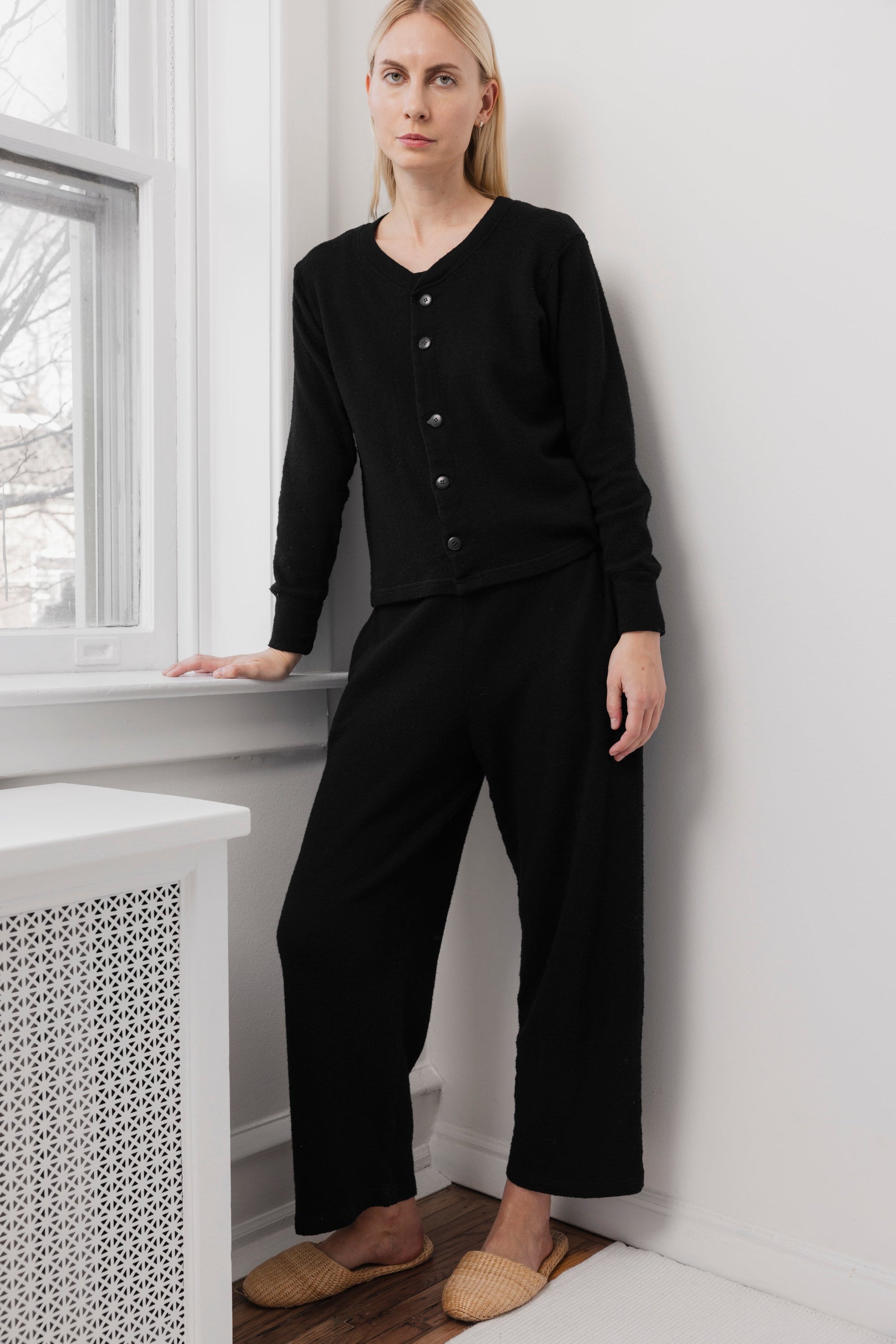 A woman with light skin and straight blonde hair stands indoors by a window, calmly looking at the camera. She wears Wol Hide's Wool Wide Leg: Onyx pants with an elastic waist, a black long-sleeve buttoned top, and beige woven slippers.
