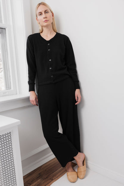 A woman with long blonde hair stands indoors by a window, wearing Wol Hide's Wool Liner in Onyx, paired with black pants and beige woven slippers. The setting features white walls and a wooden floor.