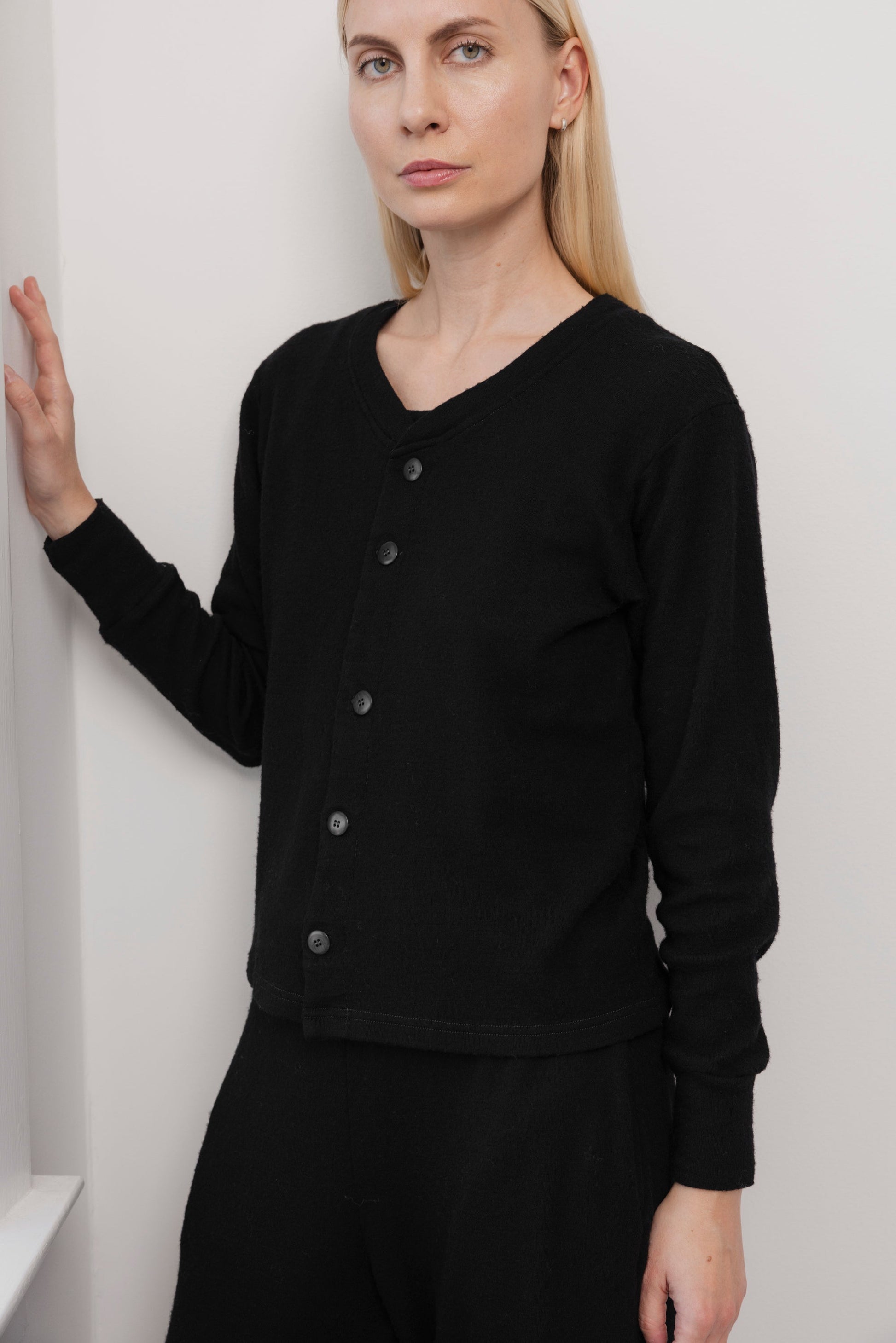 A woman with straight blonde hair wears the Wol Hide Wool Liner in Onyx, a slim black cardigan with Corozo buttons, paired with black pants. She stands indoors by a white wall, resting one hand on it and looking at the camera with a neutral expression.
