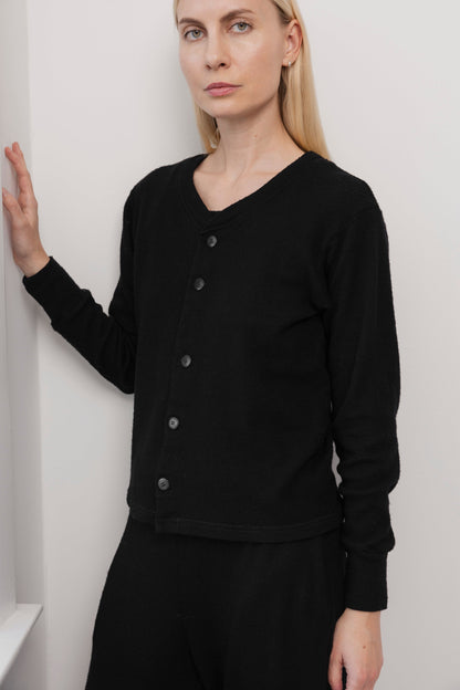 A woman with straight blonde hair wears the Wol Hide Wool Liner in Onyx, a slim black cardigan with Corozo buttons, paired with black pants. She stands indoors by a white wall, resting one hand on it and looking at the camera with a neutral expression.